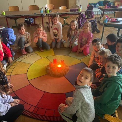 Kinder der 1a Klasse sitzen um ihren Teppich, in der Mitte brennt eine Kerze am Adventkranz